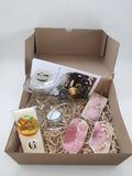 A curated gift box set containing a clear glass oil burner , pink rose and floral soaps, a tube of vanilla wax melt, and a small candle, all arranged in shredded paper inside an open brown cardboard box.