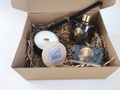 A curated gift box set containing a scented candle, a reed diffuser bottle with reeds, a small round tin, and a printed card, all arranged in shredded paper inside an open kraft cardboard box.