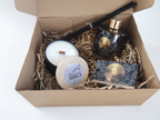 A curated gift box set containing a scented candle, a reed diffuser bottle with reeds, a small round tin, and a printed card, all arranged in shredded paper inside an open kraft cardboard box.