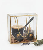 A luxury gift set in a clear acrylic and gold-framed box, containing a black scented candle with a bamboo lid, a glass diffuser bottle with a rose gold cap, and a black stick tool with a gold handle, all arranged in brown paper shred with a gold rope handle. Decorative lavender and eucalyptus sprigs sit in front on a white background.