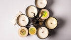 Six soy candles in black glass jars with soy wax, arranged in a circle with a glass diffuser and reeds in the center. Three of the lids are light wood with circular labels, and one label reads Gzen Candle WHITE MUSK