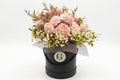 A preserved flower arrangement in a black cylindrical box with a silver monogram logo, featuring pink roses, tulips, and delicate filler flowers on a white background.