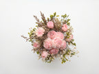 A dried flower bouquet featuring light pink roses, baby's breath, and green foliage, arranged against a plain white background.