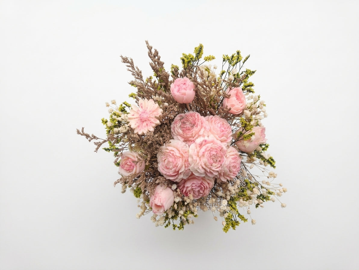 A dried flower bouquet featuring light pink roses, baby's breath, and green foliage, arranged against a plain white background.