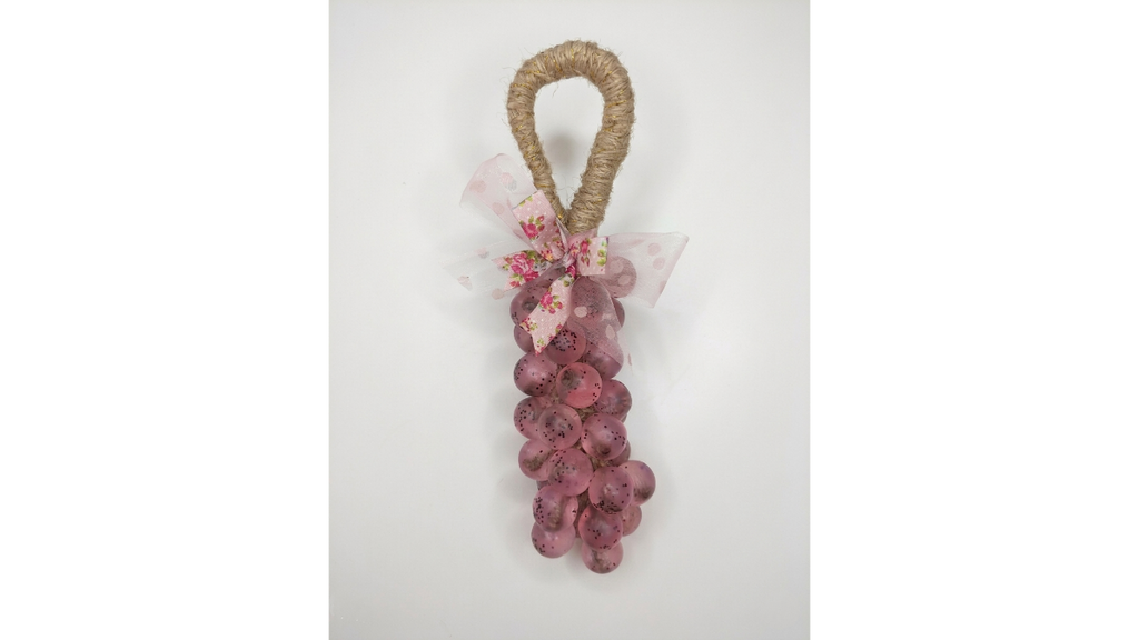 A decorative hanging ornament  handmade soap shaped like a bunch of pinkish-red grapes with a natural jute loop at the top and a sheer pink bow with small floral details, shown against a plain light background.
