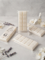 Three cream-colored soy scented wax melts chocolate bars, one in a clear plastic wrapper and two with decorative labels, arranged on a light gray surface with lavender sprigs and white flowers, by Gzen candles base in Cyprus.