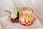 An orange and white patterned lamp jar glass candle holder filled with cream-colored wax, styled on a white marble tray with soft pink and purple flowers against a draped white fabric background. Mushroom jar by Gzen candles base in Limassol. 