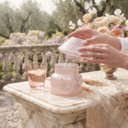 A person's hands hold a small iridescent pink glass jar lid above a matching pink glass jar on a marble table, with a pink glass and floral decor in a garden setting.