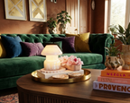 A styled living room scene featuring a deep emerald green tufted velvet sofa with accent pillows in yellow, purple, and blue. In the foreground, a round wooden coffee table holds a gold tray with a lit table lamp, pink flowers, and decorative Mushroom Lamp jar white Pure scent 