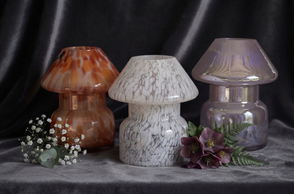Three decorative mushroom-shaped glass vases displayed on a gray fabric surface against a dark draped background. The left vase is amber-brown with orange marbling, the center vase is white with gray marbling, and the right vase is iridescent lavender-purple.