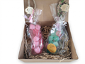A gift box containing two clear plastic-wrapped items tied with white and pink ribbons, placed in shredded paper filler inside a brown cardboard box. The left item appears pink and round, and the right item appears green and oval with a yellow label reading 'Natural Glycerin Soap Bar.