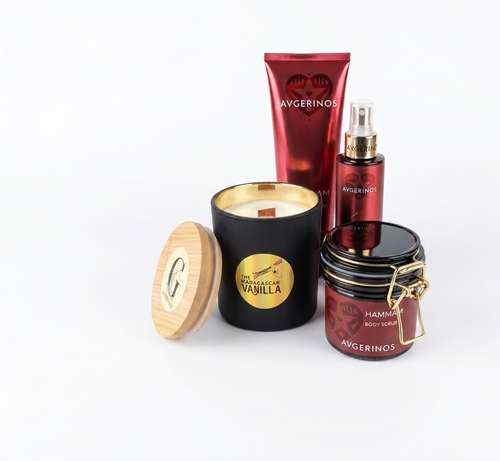 A set of four Avgerinos bath and body products arranged on a white background, including a gzen scented soy candle with a bamboo lid and wood wick,a red lotion tube hammam, a red body and hair oil spray bottle, and one jar of body scrub hammam in red and black packaging.