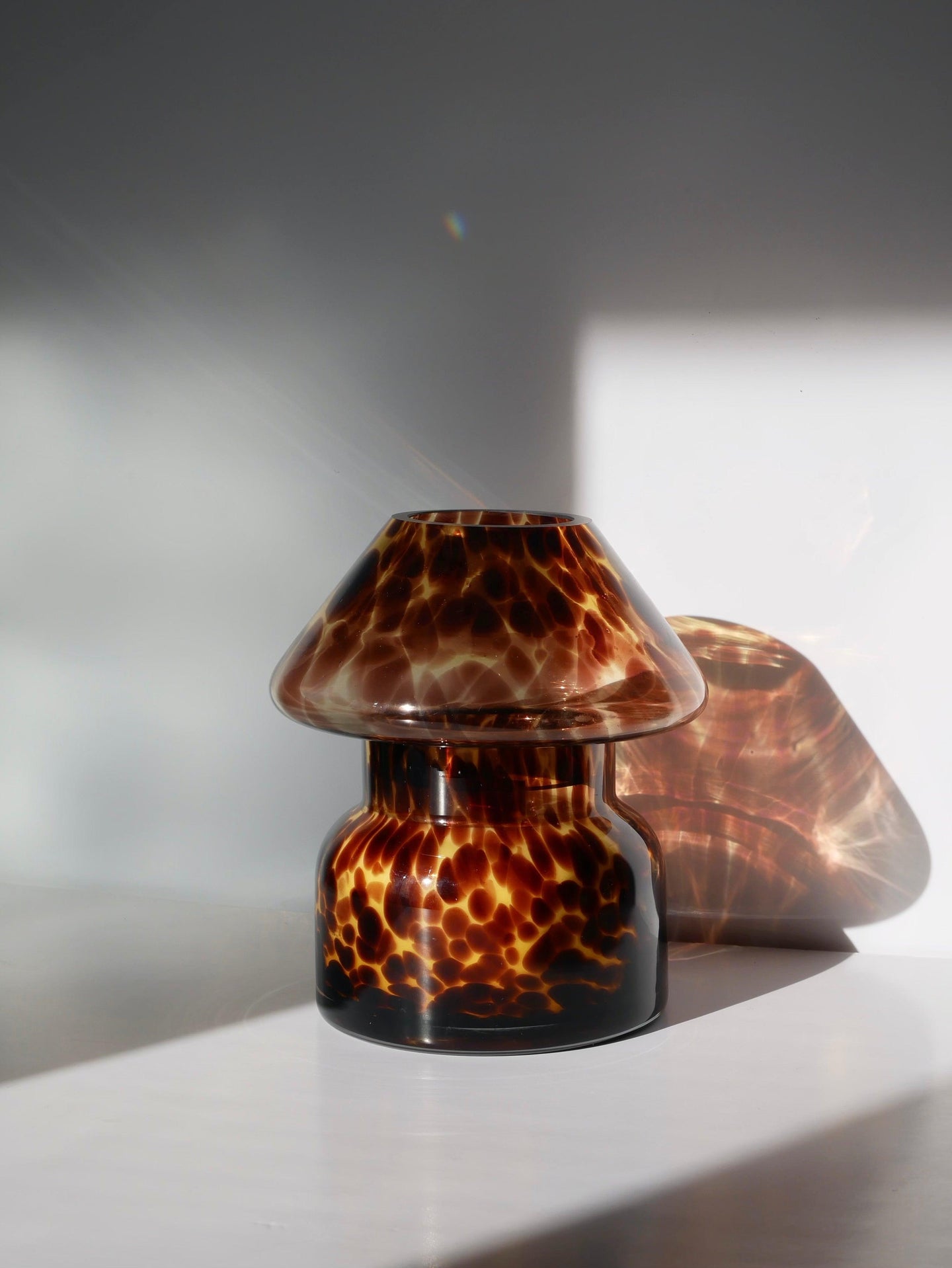 A small amber-brown glass vase with a mottled tortoiseshell pattern, shown on a white surface with dramatic sunlight and shadow.