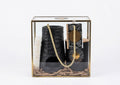 A black ceramic candle holder set inside a clear glass display box with a gold-toned frame, containing a black candle, a white candle, a gold rope, and a decorative card on a white background.