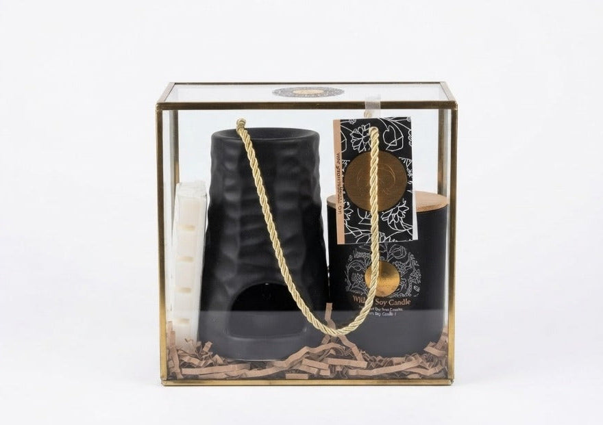 A black ceramic candle holder set inside a clear glass display box with a gold-toned frame, containing a black candle, a white candle, a gold rope, and a decorative card on a white background.