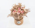 A dried flower arrangement in a round brown cardboard box, displayed on a white stool against a white background. The bouquet features light pink peony-like flowers, beige dried foliage, and small gold glitter accents.
