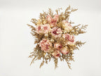 A dried floral bouquet featuring dusty pink peonies, cream carnations, and textured gold filler stems, arranged in a round bouquet against a solid white background.