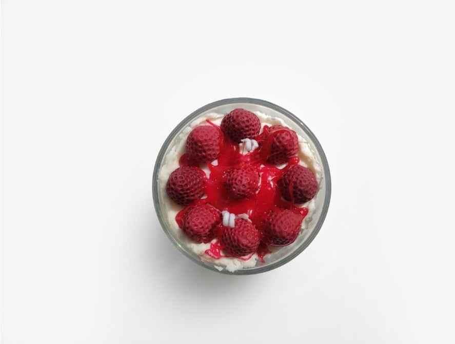 A top-down view of a clear glass candle dessert cup filled with white cream and topped with fresh red raspberries and a red berry sauce, set against a plain white background.