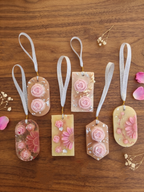 Six decorative soap wardrobe keepsake tags with white ribbon loops are arranged on a wooden surface. The tags are translucent and decorated with pink roses, daisies, and glitter, with small dried flowers and pink petals scattered around them.