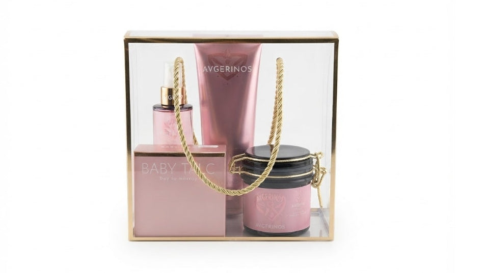 A three-piece baby talc scent gift set by Avgerinos Cosmetics in a clear acrylic box with a gold frame. The set includes a pink perfume bottle, a pink lotion tube, and a pink jar with a gold lid body oil, arranged together with a gold rope handle.