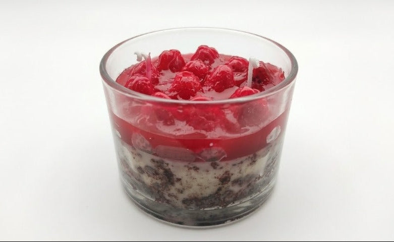 A clear glass candle jar filled with layered dark cookie crumbs, white wax, and a top layer of bright red wax and fresh raspberries, shown on a plain white background.