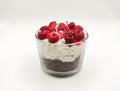 A soy candle dessert-shaped candle in a clear glass cup, featuring layers of dark chocolate, white whipped cream, and red fruit preserves topped with whole red cherries and small white wicks.