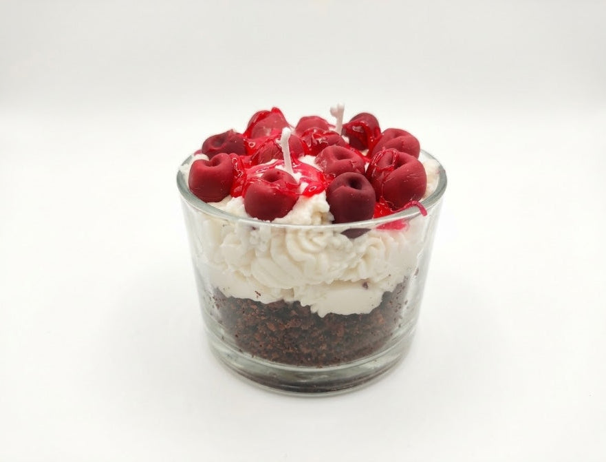 A soy candle dessert-shaped candle in a clear glass cup, featuring layers of dark chocolate, white whipped cream, and red fruit preserves topped with whole red cherries and small white wicks.