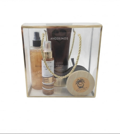 A clear acrylic gift box with a gold-toned lid and rope handle, containing several Aygerinos skincare and body care items including a dark brown tube, a spray bottle coco mist ,a coconut soy candle , set against a plain white background.