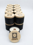 A set of black and light wood cylindrical candle tins arranged on a white background, with one tin open in front showing white wax and a wooden wick. The lids are engraved with a logo and the brand name 'OMACON WINDOWS. gzen candles cooperate gift 