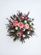 A circular floral arrangement featuring pink roses, white ranunculus, and green foliage with burgundy accents, photographed against a plain white background.