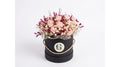 A dried flower arrangement in a round black gift box with a twine handle and a circular white emblem, photographed on a clean white background.