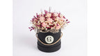 A dried flower arrangement in a round black gift box with a twine handle and a circular white emblem, photographed on a clean white background.