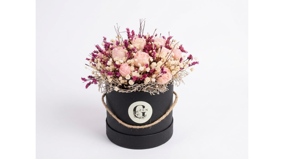 A dried flower arrangement in a round black gift box with a twine handle and a circular white emblem, photographed on a clean white background.