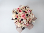 A decorative round black gift box filled with dried flowers, small pink and cream wax roses, black feather-like accents, sheer brown mesh, and a central pale heart ornament, photographed on a light gray studio background. This is a wax flower arrangement handmade by gzen candles base in Cyprus. 