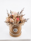 A round kraft paper box filled with dried flowers and botanicals, decorated with a pearl bead garland and a circular white logo sticker that reads 'Glen'. The box sits on a white surface against a clean white background.