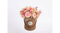 A round brown gift box filled with a bouquet of pink and beige dried-style flowers, photographed against a plain light gray background. The front of the box has a circular white label with a stylized letter G.