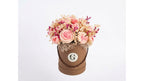 A round brown gift box filled with a bouquet of pink and beige dried-style flowers, photographed against a plain light gray background. The front of the box has a circular white label with a stylized letter G.