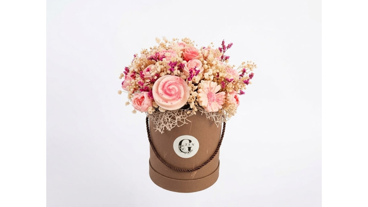 A round brown gift box filled with a bouquet of pink and beige dried-style flowers, photographed against a plain light gray background. The front of the box has a circular white label with a stylized letter G.