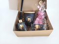 A curated gift set in an open kraft cardboard box, featuring a black reed diffuser bottle with a gold cap, a black candle tin with a gold lid, a pink wrapped plush toy, and a small packaged item with a gold medallion, all arranged on shredded paper.