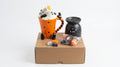 A Halloween-themed hot chocolate mug gift set displayed on a brown cardboard box against a white background. The set includes an orange mug with a tiger-striped handle and feet, a black wax warmer, small orange and black chocolate pieces, and a ghost decoration.