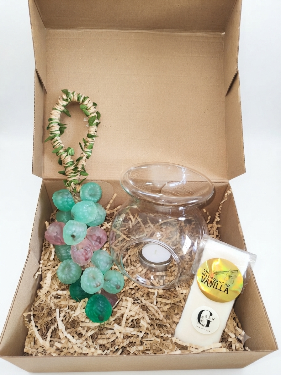 A gift box set containing a clear glass candle- oil burner with a tealight, a white vanilla-scented wax melt candle in a paper sleeve, a green and white woven garland, and a cluster of handmade natural berries soap on a bed of shredded paper inside a brown cardboard box.