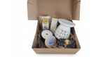 An open brown cardboard gift box filled with spa or self-care items on a white background. The contents include a white ceramic diffuser, a white candle, a packaged soap or wax item labeled 'GROOVE', a round tin labeled 'BREATHE', and a small black-and-gold card or book.