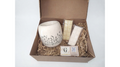 A gift set in an open brown cardboard box filled with shredded paper, featuring a speckled cream ceramic mug with black botanical line art, two cream-colored soap bars in plastic wrapping, and a small branded card. The brand name 'G' is visible on the card.