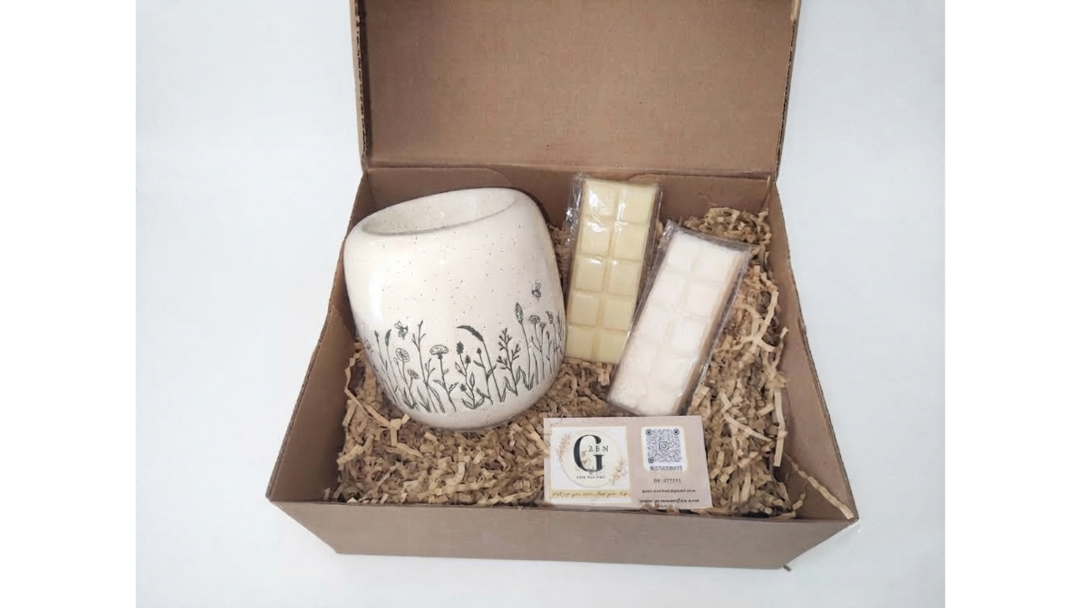 A gift set in an open brown cardboard box filled with shredded paper, featuring a speckled cream ceramic mug with black botanical line art, two cream-colored soap bars in plastic wrapping, and a small branded card. The brand name 'G' is visible on the card.