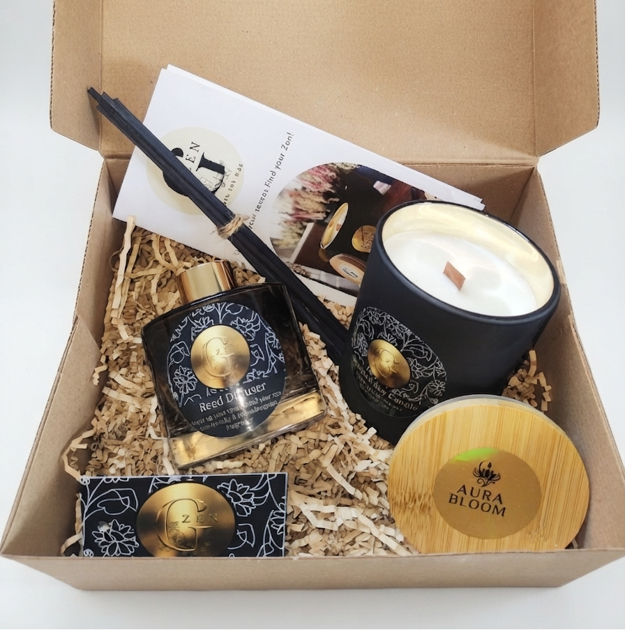 A curated gift box set from Aura Bloom, displayed inside an open brown cardboard box filled with shredded paper. The set includes a black soy candle with a bamboo lid, a black and gold perfume reed diffuser  bottle, a black and gold soap bar, a wooden diffuser wand, and a printed card.