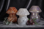 Three decorative mushroom-shaped glass vases displayed on a gray fabric surface against a dark draped background. The left vase is amber-brown with orange marbling, the center vase is white with gray marbling, and the right vase is iridescent lavender-purple. Murble jar by Gzen candle base in Limassol , Cyprus . Best gift for her or him. Handmade in Cyprus-Handpured in mushroom jar with natural ingredients eco-friendly and hypoallergenic fragrance with soy and coconut wax . 