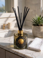 A reed diffuser bottle with black reeds and a gold cap sits on a marble bathroom counter next to a folded white towel and a potted succulent.