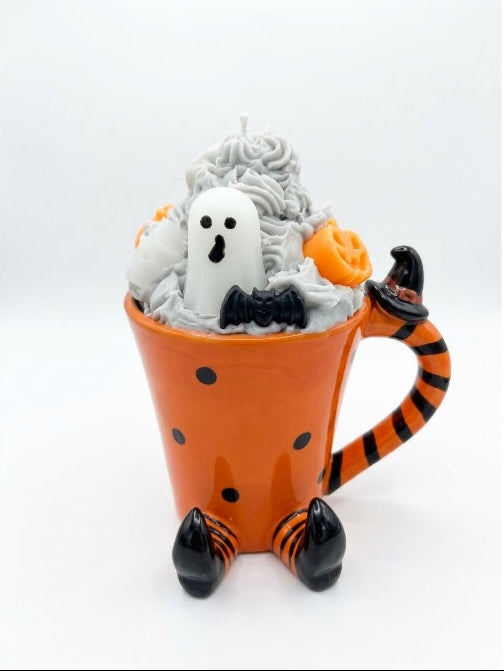 A Halloween-themed ceramic mug whipped candle with an orange body, black polka dots, and a handle shaped like a witch's tail. The top is filled with gray whipped wax, a white ghost figure, a small black bat, and orange pumpkin decorations, set against a plain white background.
