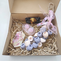 A gift box filled with bath and body items, including pink and lavender bath bombs, a pink soap bar, a small black box with a gold medallion, and a pink ribbon-tied loop, arranged in shredded paper inside an open brown cardboard box.