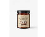 An amber glass jar of freshly baked Greek kourabiedes with a black lid and a beige label reading 'Freshly Baked Kourabiedes at Grandma's', set against a clean white background. Scented Soy Candle base in Limassol, Cyprus. Christmas Candle